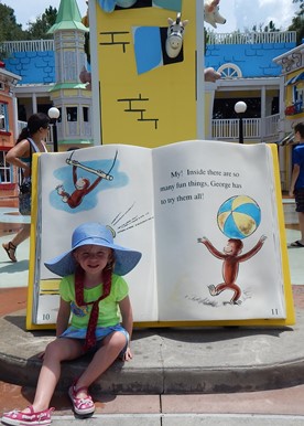 little girl in blue hat sitting with Curious George book on JAJF trip 
